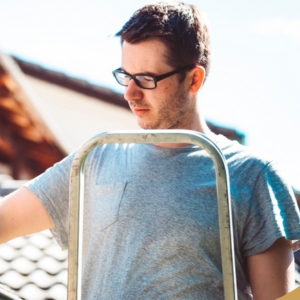 a person wearing glasses and a gray shirt, inspecting a gutter while standing on a ladder